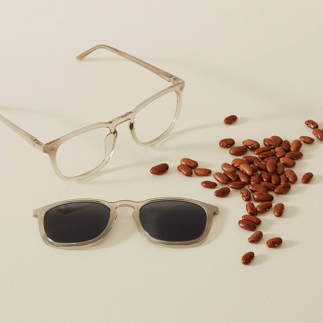 A pair of crystal coloured glasses, with a clip-on and pile of castor beans laying flat on a beige background